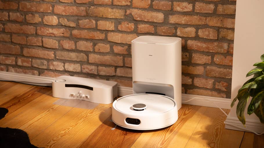 A white robot vacuum cleaner and its charging station next to a brick wall.