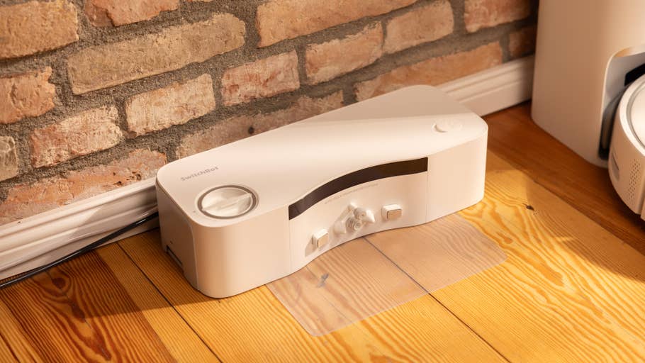 A white SwitchBot S10 device on a wooden floor in front of a brick wall.