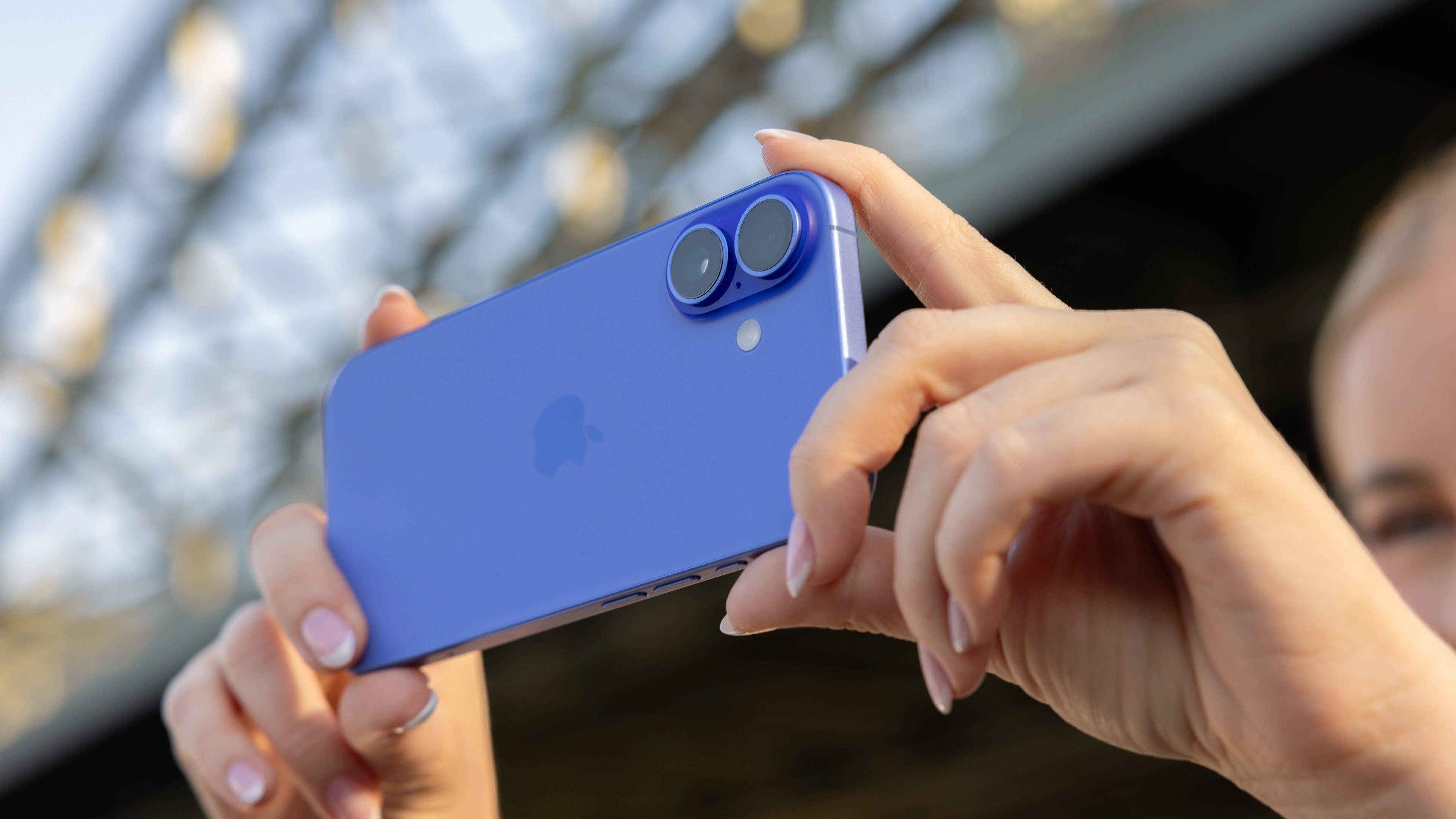 A person holding a blue iPhone 16 with dual cameras in a sunny outdoor setting. A person holding a blue iPhone 16 with dual cameras in a sunny outdoor setting.