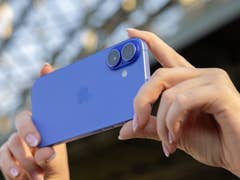 A person holding a blue iPhone 16 with dual cameras in a sunny outdoor setting.