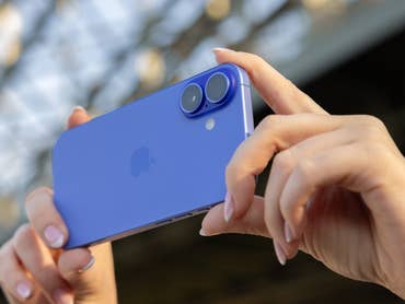 A person holding a blue iPhone 16 with dual cameras in a sunny outdoor setting.