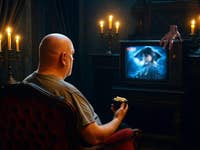A bald man sitting in an armchair, watching a spooky scene on an old TV, with candles illuminating the dark room.