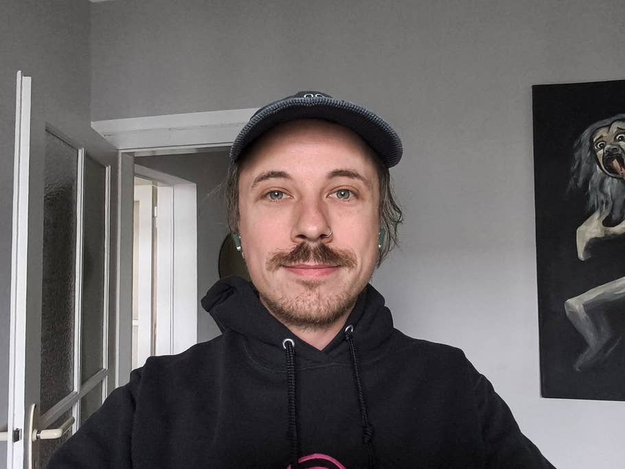 A young man with a mustache and a cap smiling in a room with a painting on the wall.