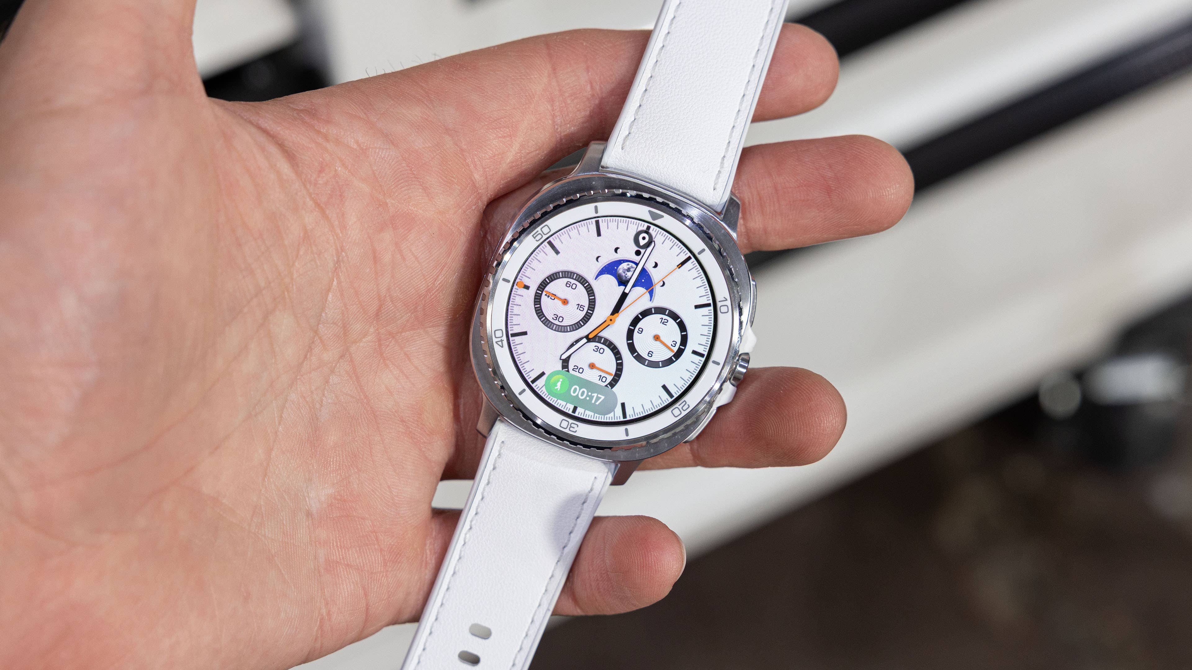 A person holding a Samsung Galaxy Watch 8 Classic with a white strap, displaying various watch faces.
