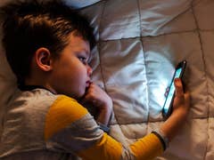 A child lying on a bed, looking at a smartphone screen in the dim light.