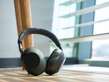A pair of Sony WH-1000XM6 headphones lies on a wooden floor next to a window.