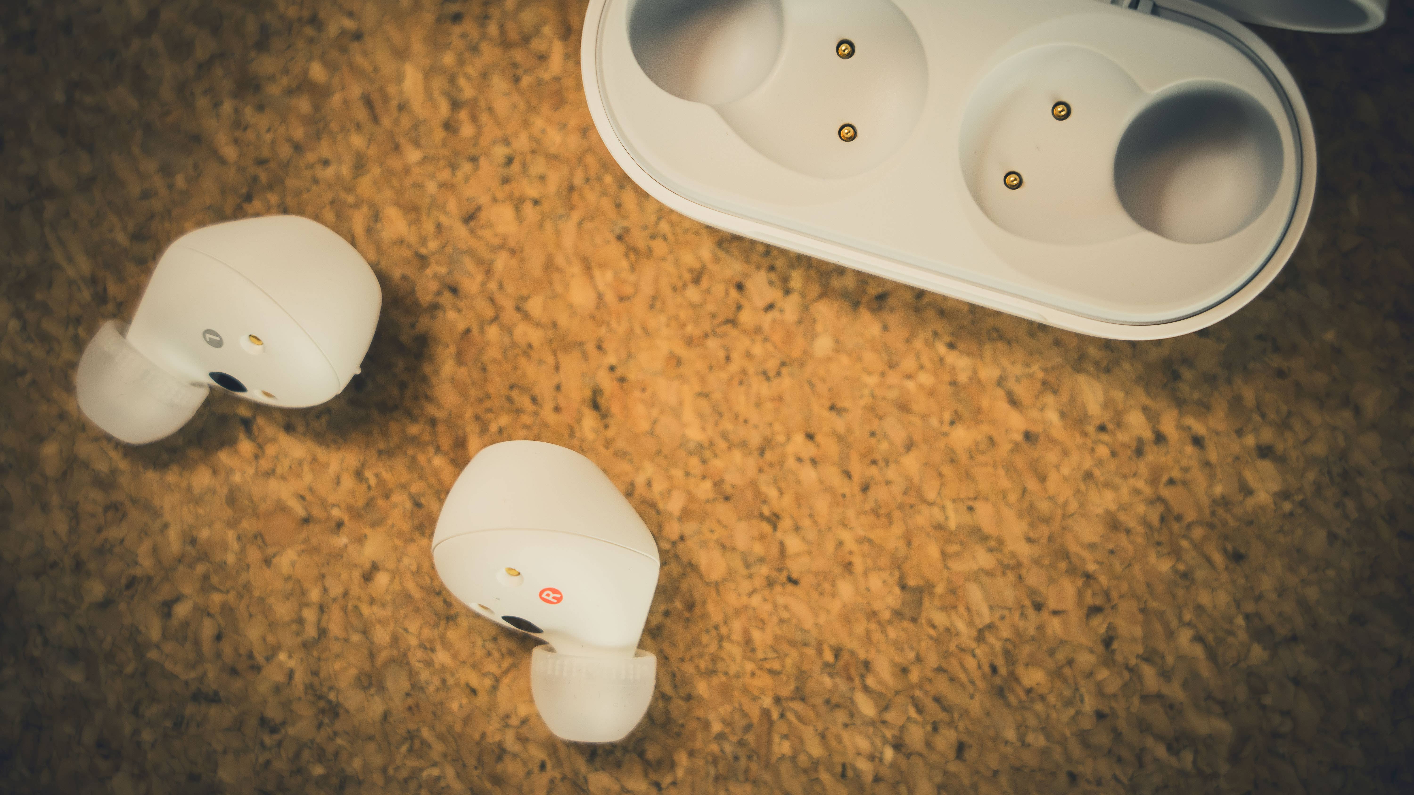 White Sony wireless earbuds next to their charging case on a cork surface.