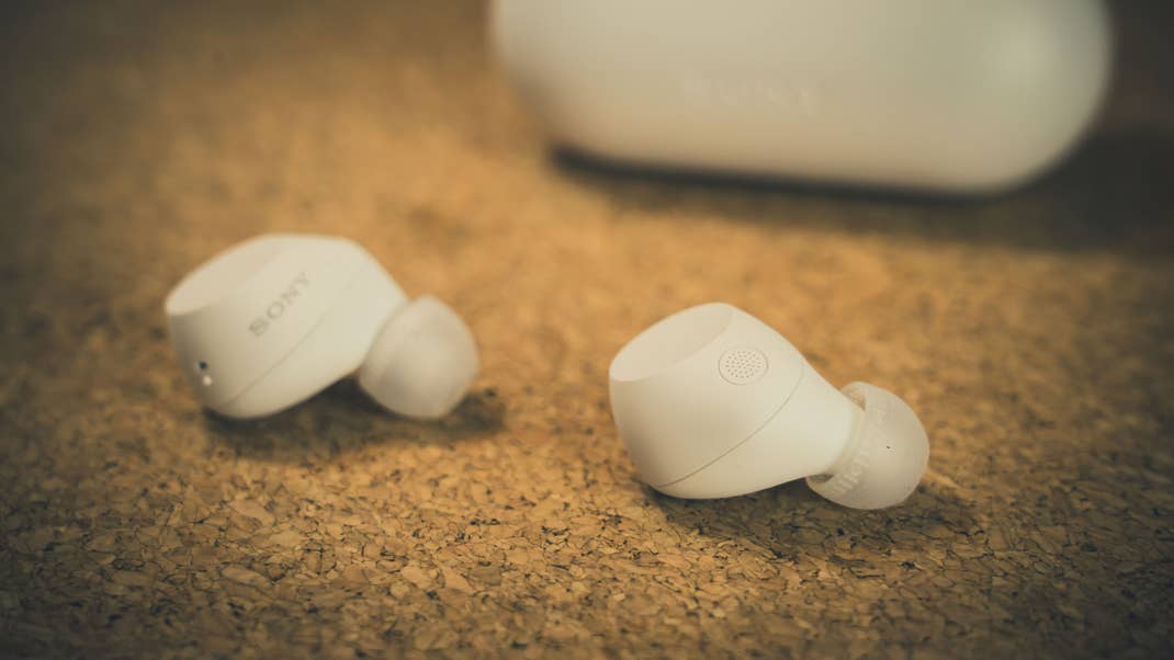 White Sony earbuds on a cork surface with a charging case in the background. White Sony earbuds on a cork surface with a charging case in the background.