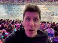 A man takes a selfie in a crowded arena during a swimming event, with colorful lights in the audience.