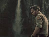 A man standing near a waterfall, wearing a beige shirt, with a rocky cliff in the background.