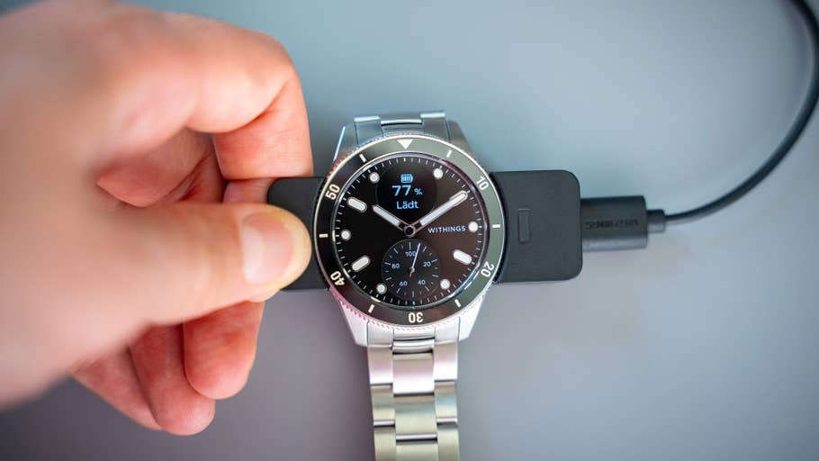 A hand holds a Withings ScanWatch Nova while charging on a docking station, showing 77% battery.