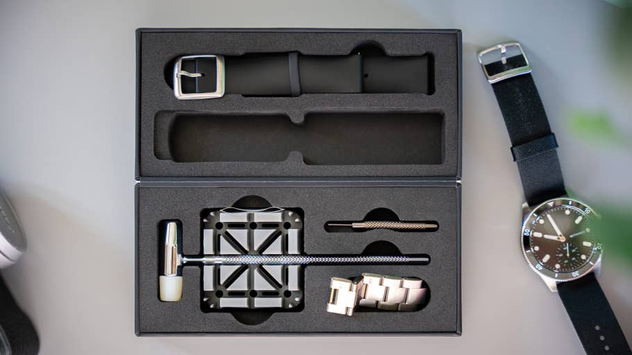 An open box with a black watch strap, tools and a silver watch.